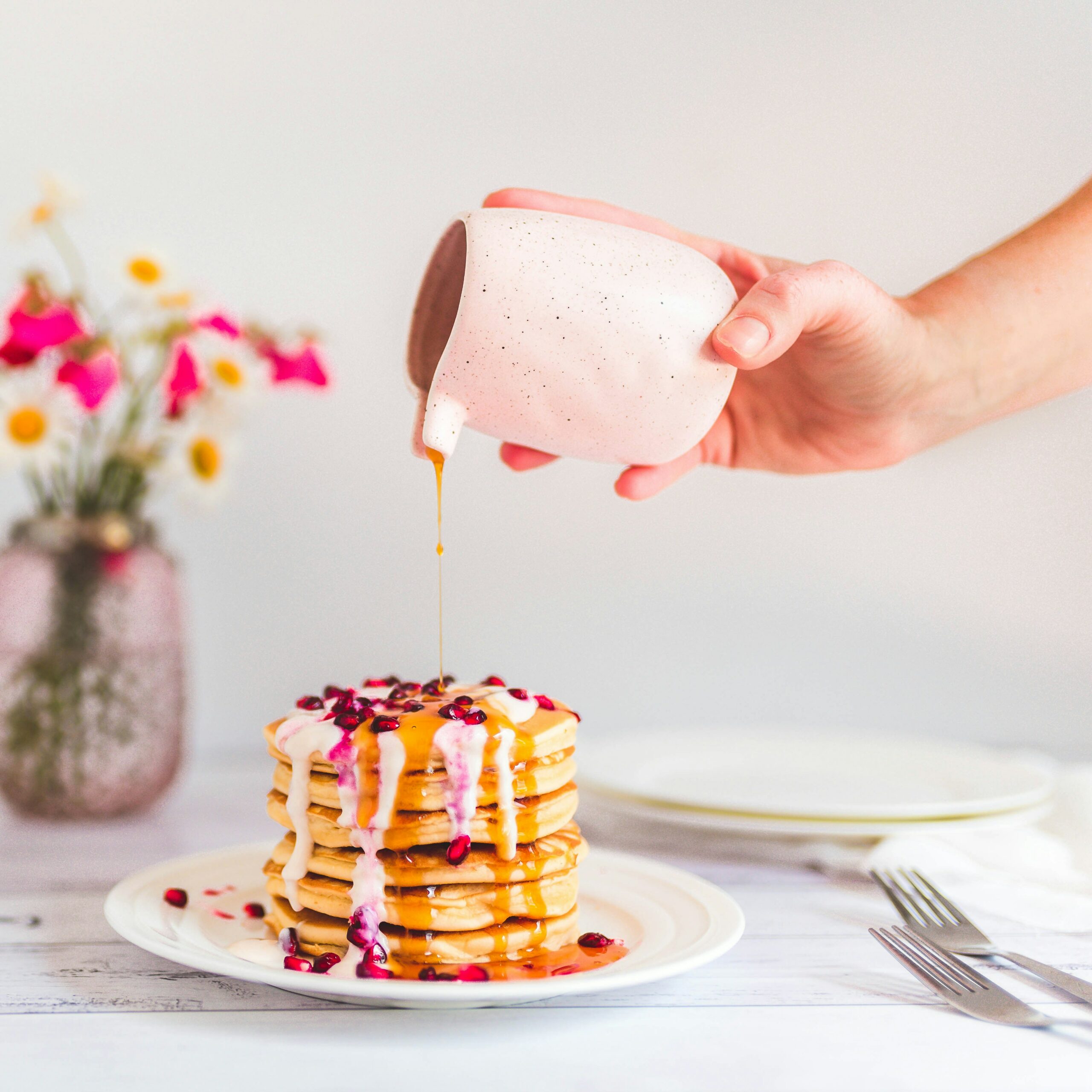 A pink ceramic pot is pouring maple syrup onto a stack of golden pancakes on a white plate. A bouquet of pink, white and yellow flowers is in the background.