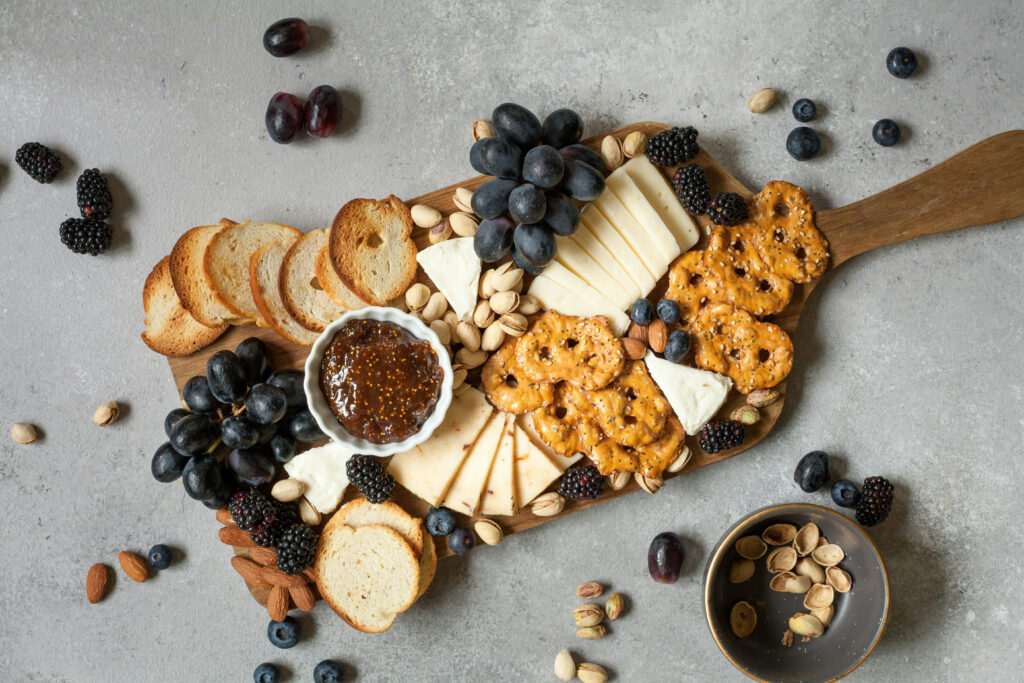 A cutting board with toasted crackers, pretzel chips, purple grapes, blackberries, blueberries, almonds, pistachio, fig jam, brie cheese, pepperjack cheese and cheddar cheese. A small bowl for pistachio shells. This cheese board is an example of a healthy and balanced snack option.